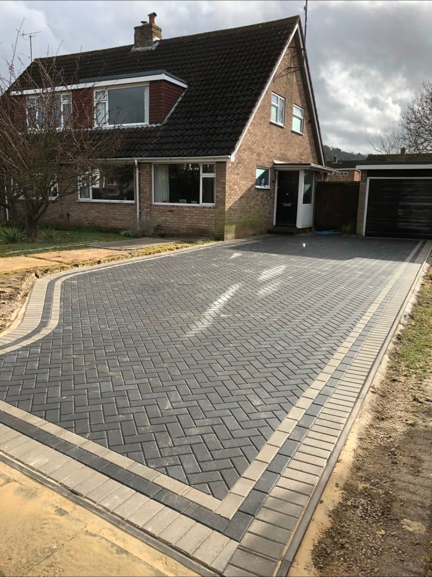 Dark grey herringbone block paving driveway with light grey decorative border in front of house.