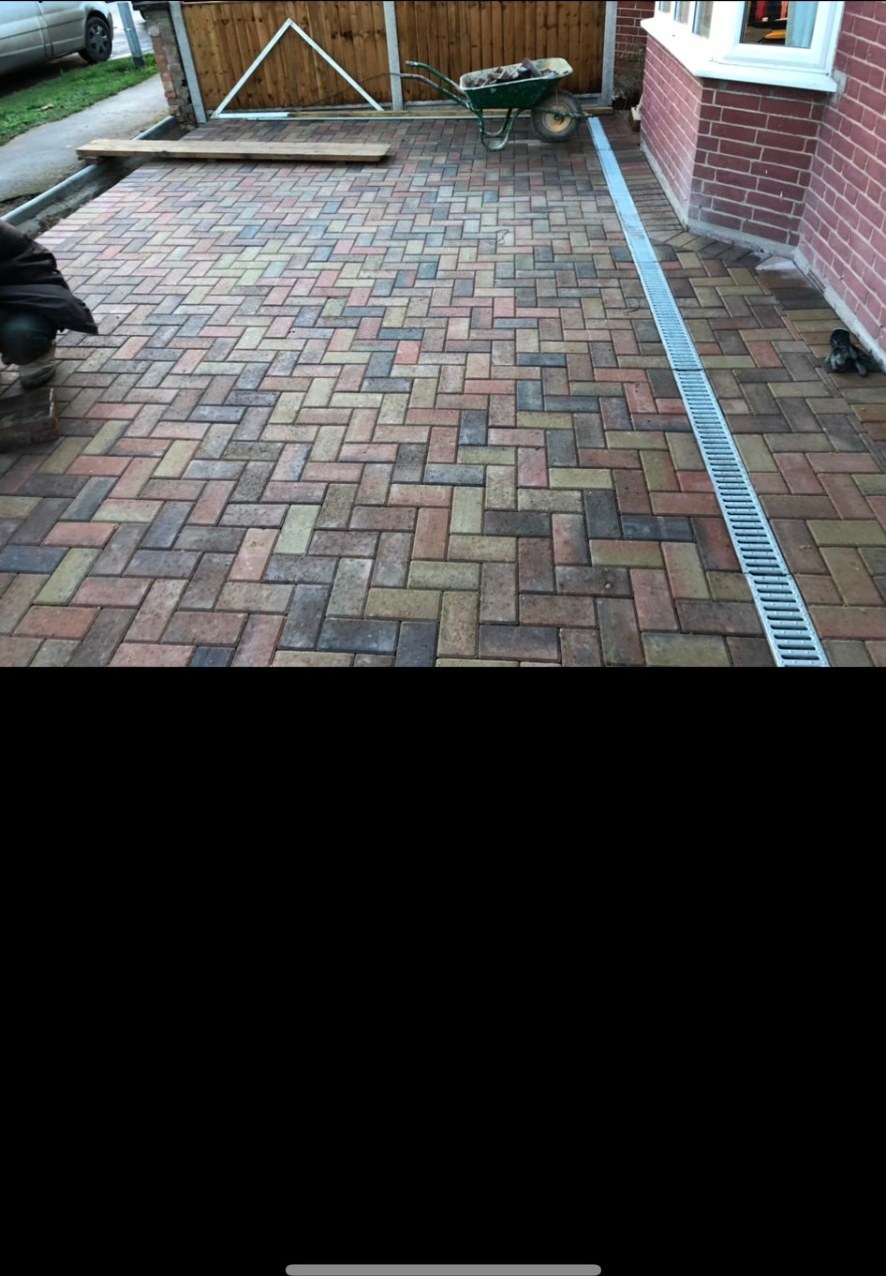 Herringbone pattern block paving driveway with linear drainage channel by a brick house.