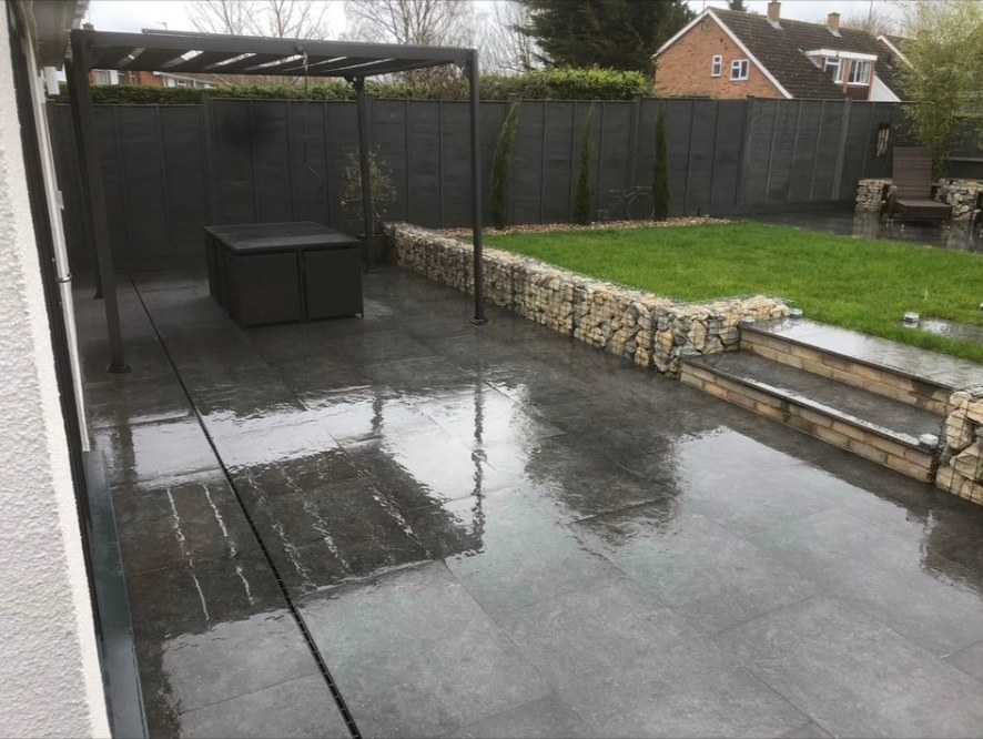 Dark porcelain patio with gabion stone retaining wall, lawn, and modern pergola.